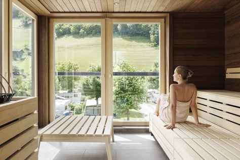 Frau im Bademantel in der Sauna Das Innere der Sauna gebaut aus Holz mit riesigen Fenstern, eine Frau im Bademantel sitzt auf der Bank, mit Blick auf die schöne grüne Landschaft